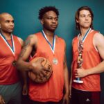 Three basketball players celebrating with medals and a trophy in victory pose.