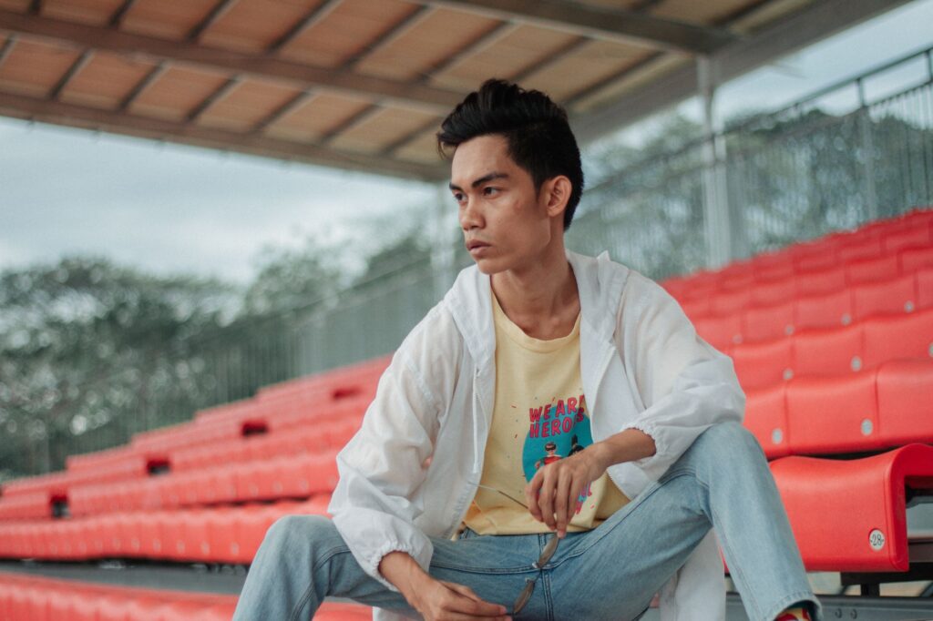 Young man posing casually on red stadium seats wearing denim jeans and a white jacket.