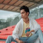 Young man posing casually on red stadium seats wearing denim jeans and a white jacket.