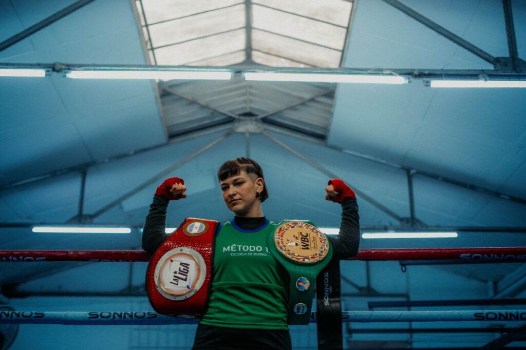 A victorious boxer poses triumphantly with championship belts in an indoor gym.