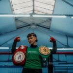 A victorious boxer poses triumphantly with championship belts in an indoor gym.
