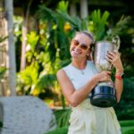A woman wearing sunglasses holds a trophy, smiling in a lush tropical garden setting.