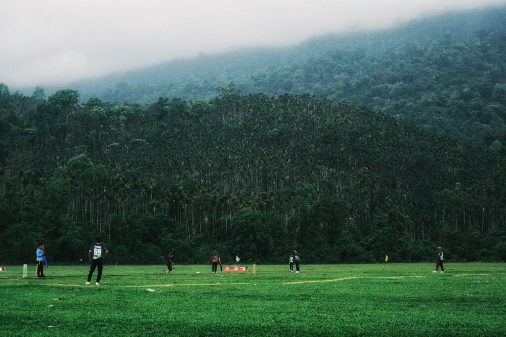 Cricketers playing in a lush green field surrounded by a forest in Sylhet, Bangladesh.