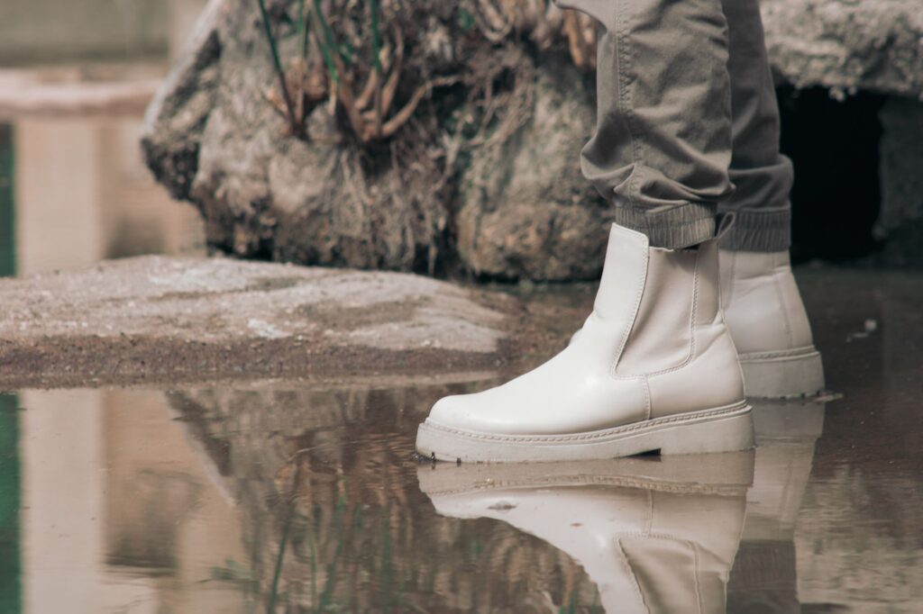 White Chelsea boots in a puddle with a rocky outdoor background, showcasing a stylish and reflective moment.