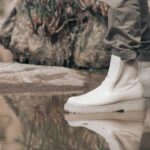 White Chelsea boots in a puddle with a rocky outdoor background, showcasing a stylish and reflective moment.