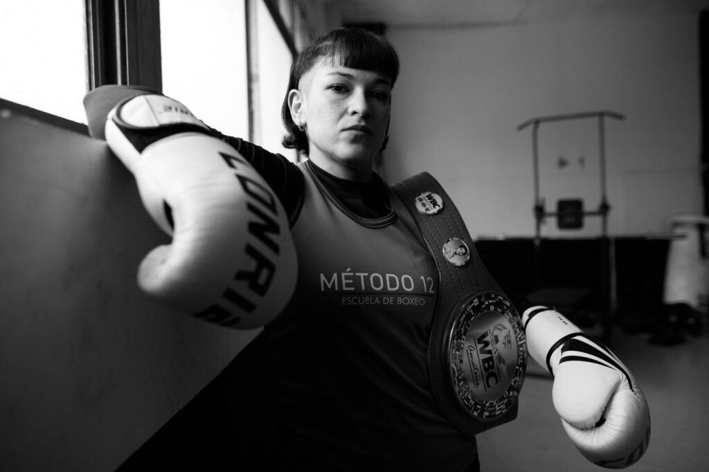 Black and white portrait of a female boxer with gloves and championship belt in a Buenos Aires gym.