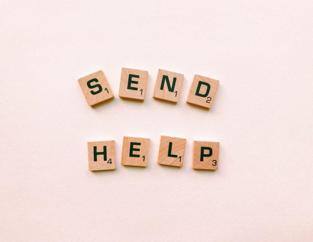 Wooden Scrabble tiles arranged to form the phrase 'send help' on a light pink background.