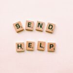 Wooden Scrabble tiles arranged to form the phrase 'send help' on a light pink background.