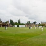 Outdoor cricket match on a lush green field with a historic building backdrop, overcast sky, many players visible.