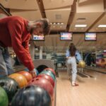 A man in a red sweater selects a bowling ball while a woman rolls a ball down the lane.