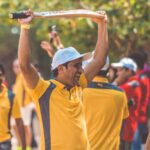 A cricketer in yellow uniform celebrating with a cricket bat raised in triumph outdoors.