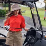 Woman in red and khaki golfing outfit standing by a cart, holding a golf club on a sunny day.