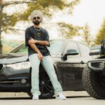 Man with turban and sunglasses striking a pose between a BMW and Jeep outdoors.