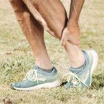 Man holding his calf in pain, wearing teal sneakers on green grass.