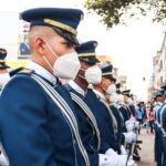 Soldiers in ceremonial uniforms wearing face masks marching in a parade on a street in Ecuador.