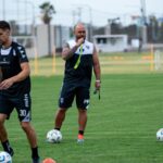 Focused soccer coach instructing players during a training session on a green field.