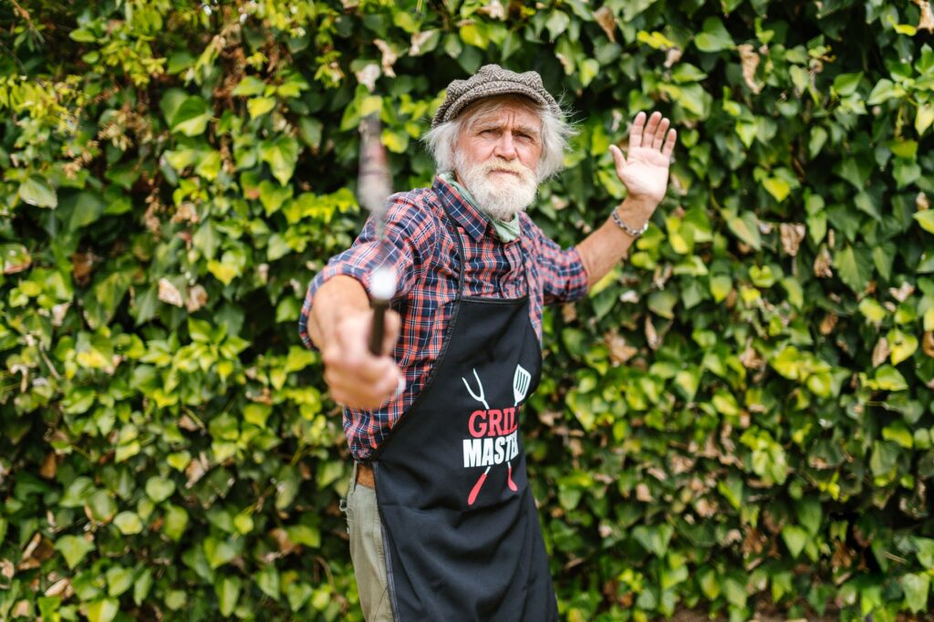 Elderly man wearing a grill master apron holds a spatula outdoors against a leafy background.