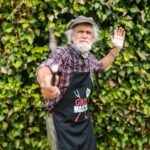Elderly man wearing a grill master apron holds a spatula outdoors against a leafy background.