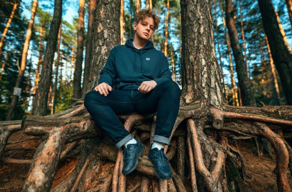 A young adult man sits on large tree roots in a sunlit forest, wearing casual clothing.