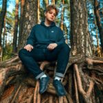 A young adult man sits on large tree roots in a sunlit forest, wearing casual clothing.