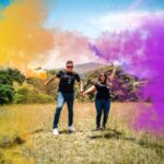 A joyful couple holding smoke bombs in a vibrant open field, celebrating together.