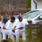 Group of cricket players sitting on a bench outdoors, dressed in white uniforms.