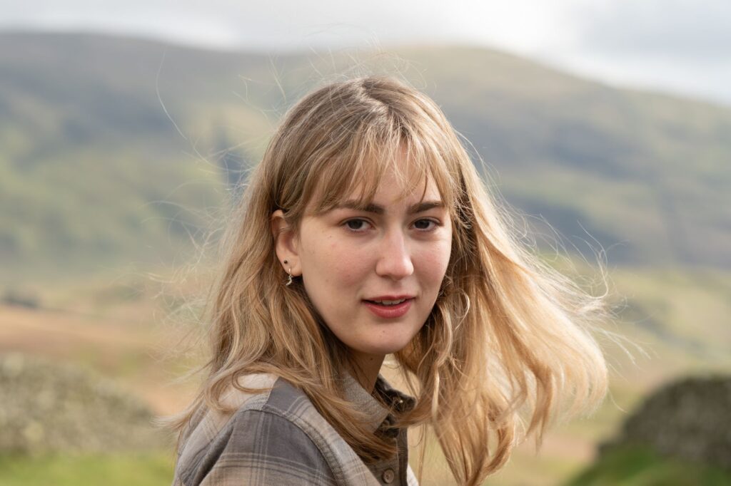 A woman with blonde hair poses in the windy hills of Windermere, England.