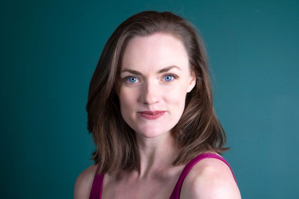 Portrait of a confident woman with blue eyes and brown hair against a teal background.