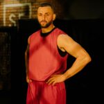 Bearded man in red activewear stands confidently with hands on waist indoors, looking determined.