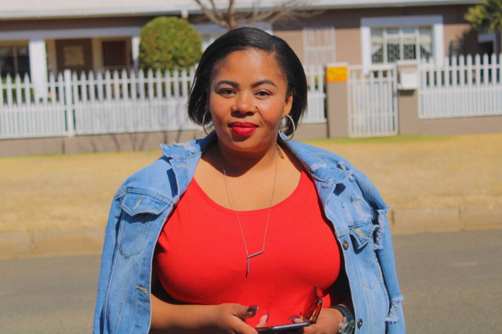 African woman in a red dress and denim jacket confidently holding her smartphone outdoors.