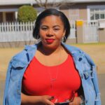 African woman in a red dress and denim jacket confidently holding her smartphone outdoors.