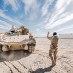 Bradley Fighting Vehicle and soldier in desert training operation under clear sky.