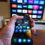 Close-up of a hand holding a phone displaying streaming apps in front of a TV with multiple app icons.