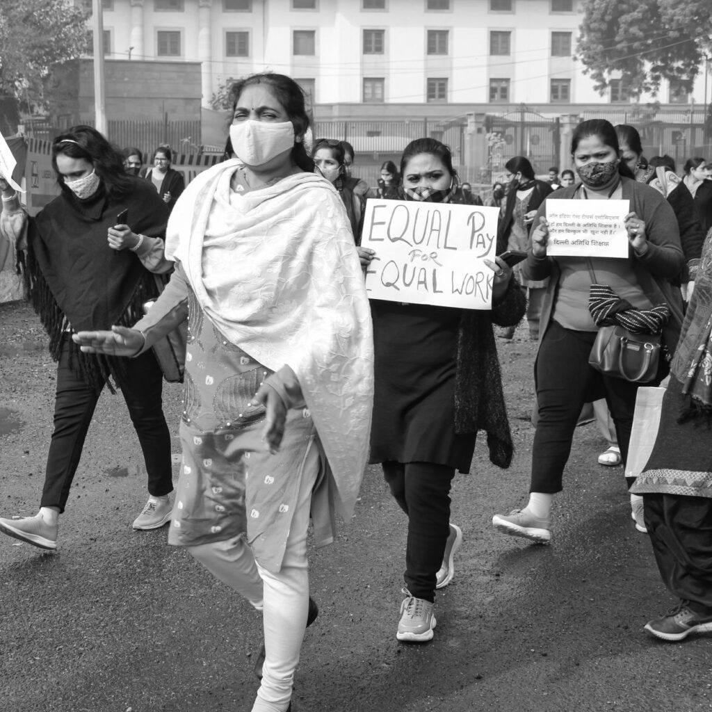 A black and white image of a women's rally advocating for equal pay, with participants wearing face masks and holding placards.