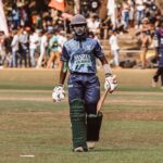 Cricketer wearing protective gear with bat in hand during a match. Crowd and teammates in background.