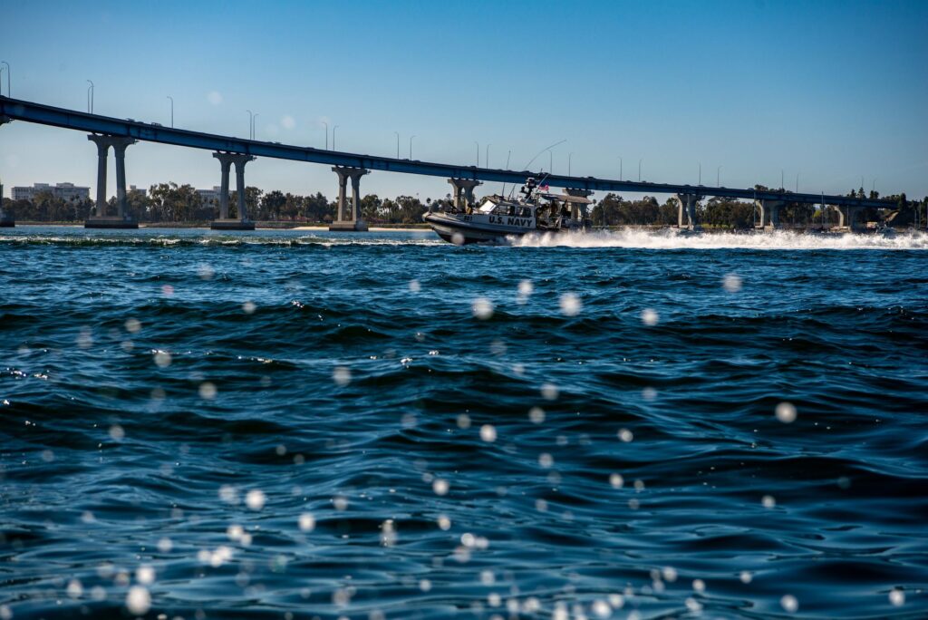 Navy patrol boat speeding under a bridge, creating waves and ripples in the water.