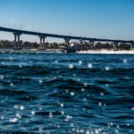 Navy patrol boat speeding under a bridge, creating waves and ripples in the water.
