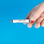Gloved hand holding COVID-19 test tube with blood sample against blue background.