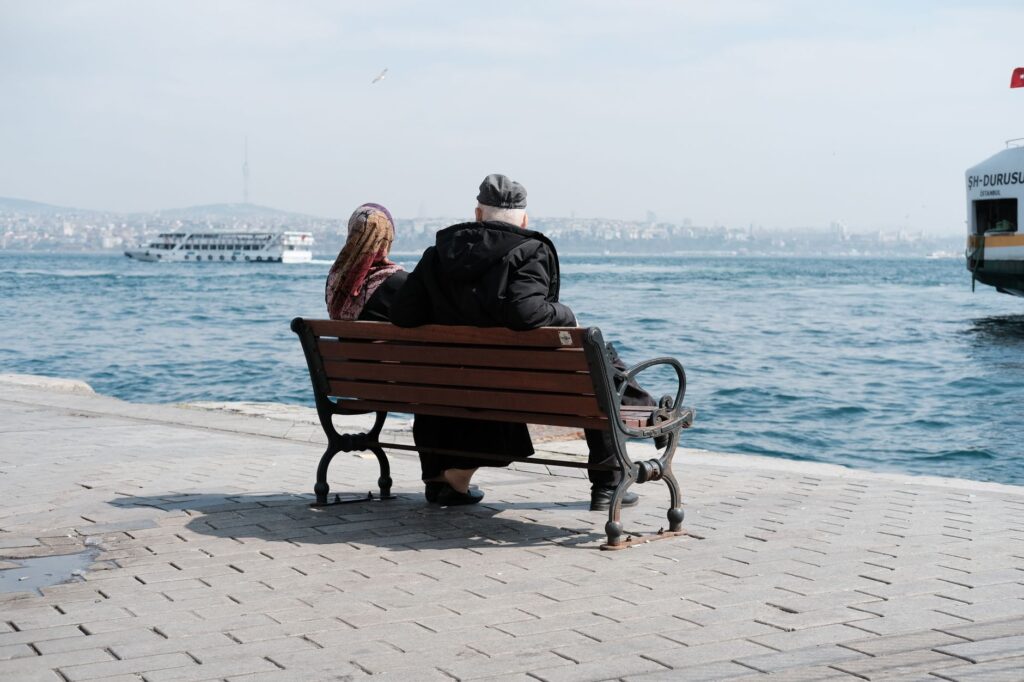 Senior couple sitting on a bench by the sea, with cityscape and ferry in view.