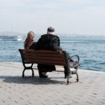 Senior couple sitting on a bench by the sea, with cityscape and ferry in view.