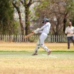 Batsman in white gear hits cricket ball on sunny outdoor field.