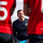 A soccer coach discusses strategy with players during daytime practice outdoors.