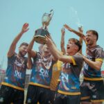 A joyous soccer team celebrates with a trophy outdoors under a clear sky.