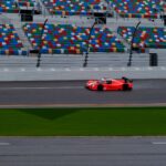 A vibrant red race car speeding on a track with empty colorful stands in the background.