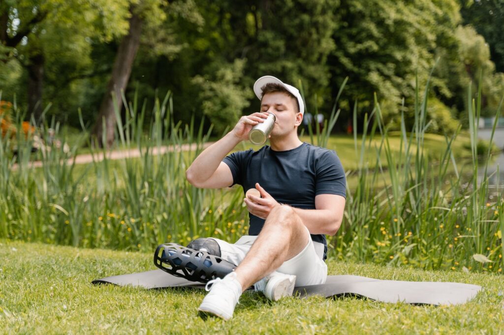 A man with a prosthetic leg sitting on a yoga mat outdoors, drinking from a tumbler.