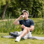 A man with a prosthetic leg sitting on a yoga mat outdoors, drinking from a tumbler.