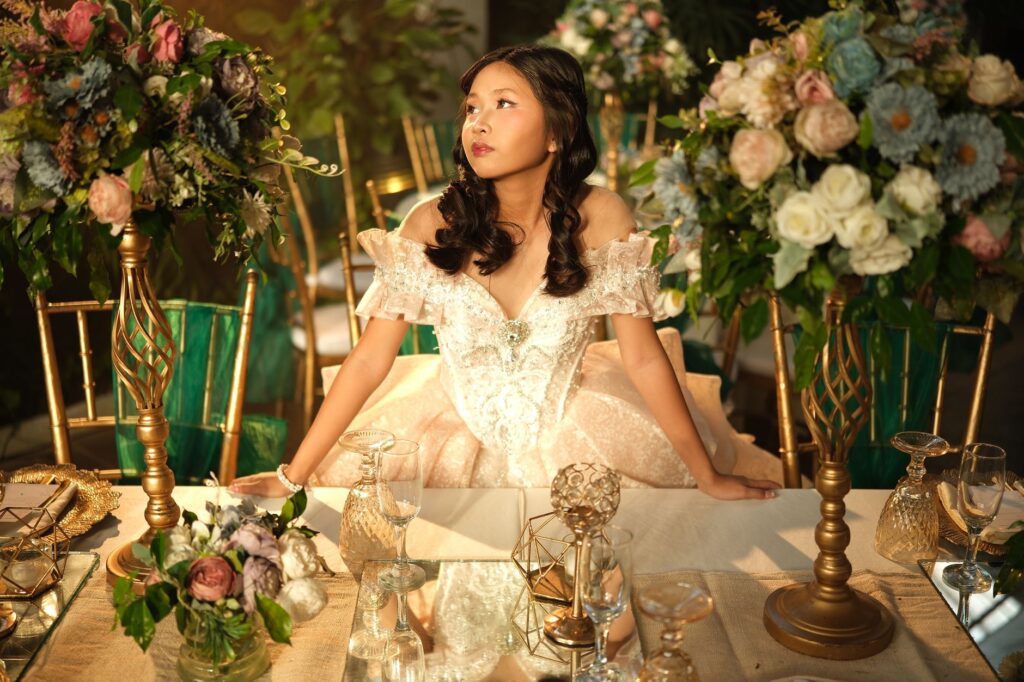 A woman in a formal gown poses elegantly at a lavishly decorated table with floral arrangements.
