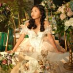 A woman in a formal gown poses elegantly at a lavishly decorated table with floral arrangements.