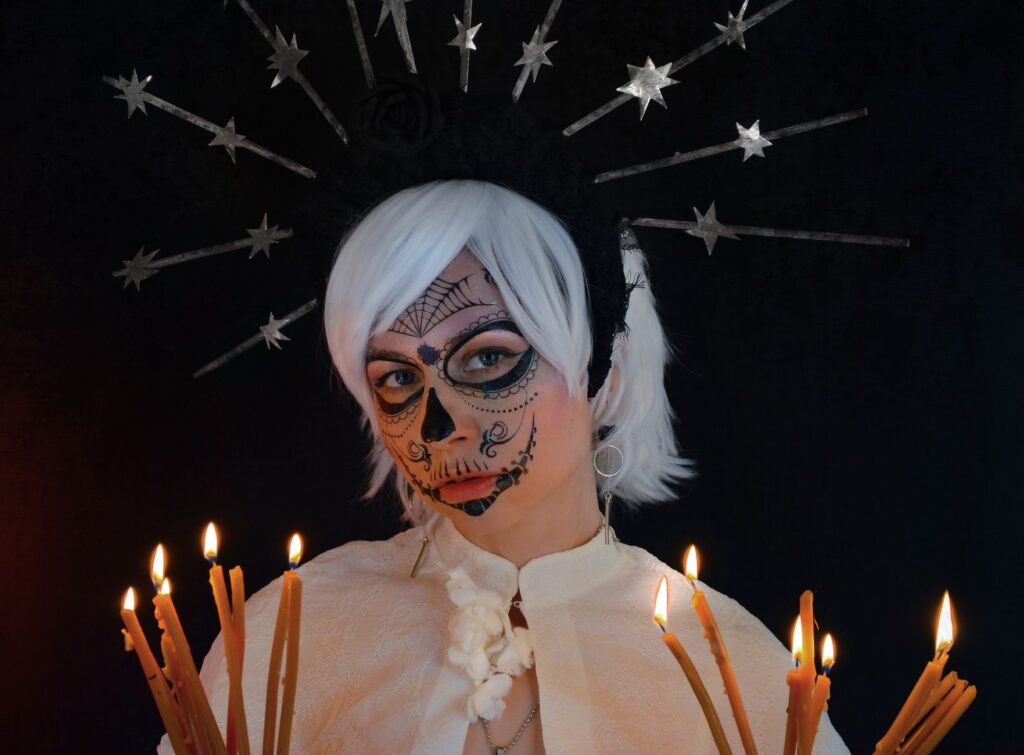 Concentrated young female model with creative Mexican styled makeup and headband standing against black background with burning candles and looking at camera during Dia de los Muertos celebration