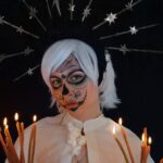Concentrated young female model with creative Mexican styled makeup and headband standing against black background with burning candles and looking at camera during Dia de los Muertos celebration